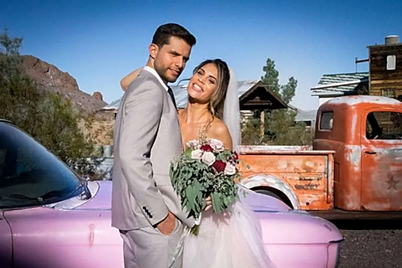 Un jeune couple marié se tient devant une vielle cadillac rose et un camion ancien dans un village fantôme abandonné